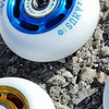 Four inline skate wheels with different colored hubs (black, blue, red) and bearings visible, lying on a gravel surface. 